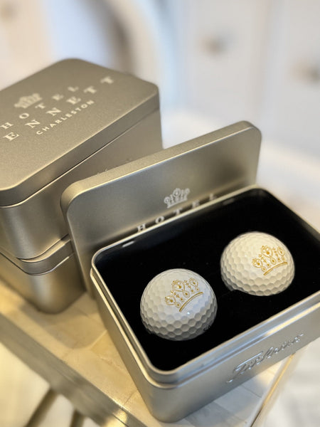 Two golf balls with a crown emblem in an open silver tin on a blurred background
